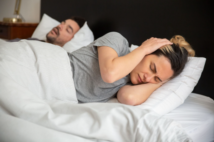 A person lies awake in bed pressing a hand to the side of their head while another person sleeps beside them.