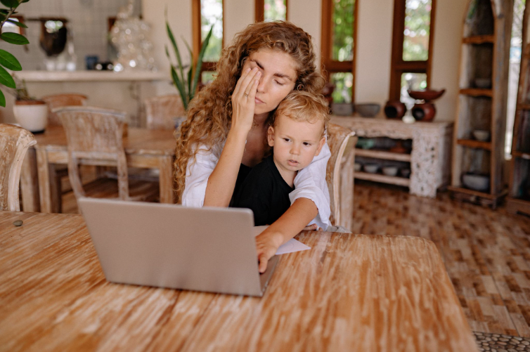 A mom rubs her eye while working on a laptop with a toddler on her lap.