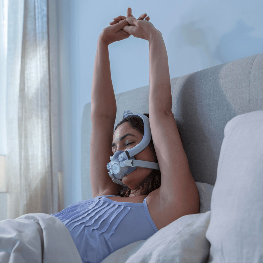Woman in bed stretches after a good night of sleep while still wearing her AirTouch F30i Comfort full face CPAP mask